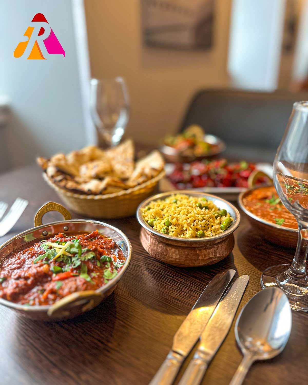 Indian meal with curry, yellow rice, naan, and sides on a wooden table with glasses and cutlery. 